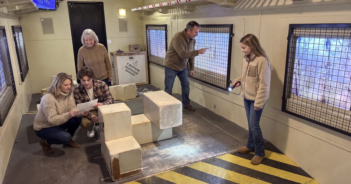 A family in fall sweaters solves a paper clue among wooden crates inside the Runaway Train escape room car while a girl shines a flashlight at the window